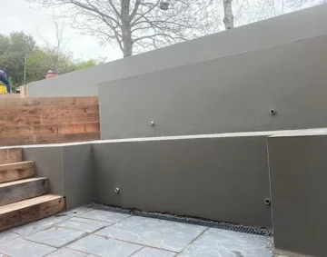 A modern outdoor terrace featuring smooth grey rendered retaining walls with small circular fittings. Wooden steps lead upward on the left, and slate tiles form the lower patio area.