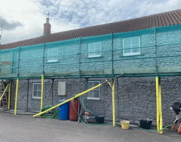 A two‑storey stone building with a tiled roof is surrounded by scaffolding covered in turquoise safety netting. Yellow poles support the structure, and various construction materials. Hoses, barrels and buckets are scattered along the paved ground below. The scene shows renovation work in progress under a partly cloudy sky.