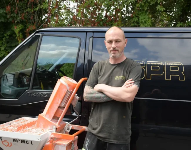 Steve Sheppard stands confidently beside his black van bearing the gold “SPR” (Sheppard Plastering Rendering) logo. To his side is an orange plastering machine.