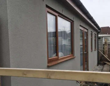 Side view of a modern single-storey building with smooth grey render and wooden-framed windows and doors, showcasing a clean finish and attention to detail in exterior rendering work.