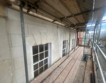 Scaffolding platform along the upper floor of a row of buildings, showing light stone façade with decorative window arches and white-framed windows, part of an exterior restoration or rendering project overlooking a street lined with colourful houses.