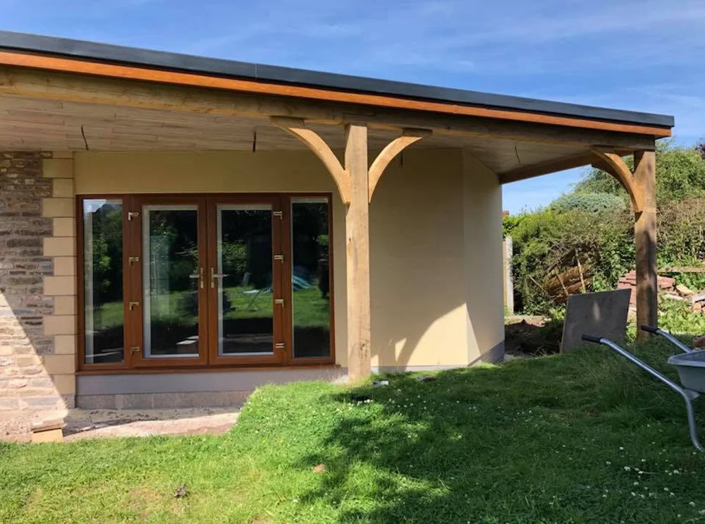 Exterior view of a modern house extension with smooth cream render, wooden beams supporting a covered patio roof, and large glass doors framed in wood, showcasing quality craftsmanship and clean architectural design.