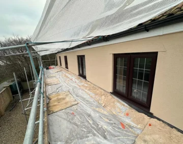 Exterior view of a cream-coloured rendered wall with dark-framed windows, seen from scaffolding covered with protective sheeting during renovation work, showcasing smooth render finish and careful preparation.