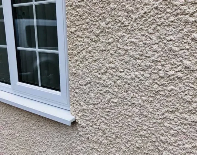 Close-up of a cream-coloured exterior wall with a roughcast render finish beside a white-framed window, showcasing the smooth edges and textured surface typical of a high-quality rendering service.